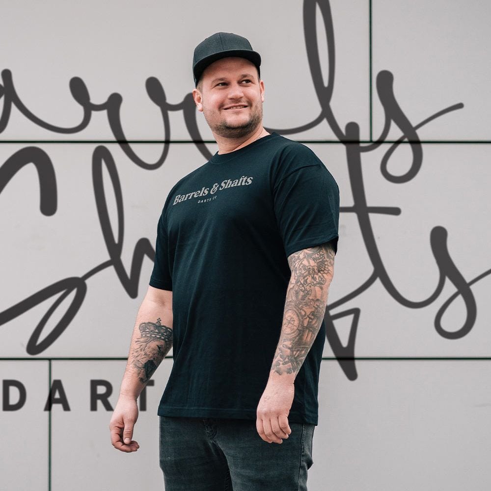 Barrels and Shafts T-Shirt - Black Ein Mann trägt ein schwarzes T-Shirt mit der Aufschrift „Barrels & Shafts“. Er steht vor einer Wand mit passender Schrift im Hintergrund.