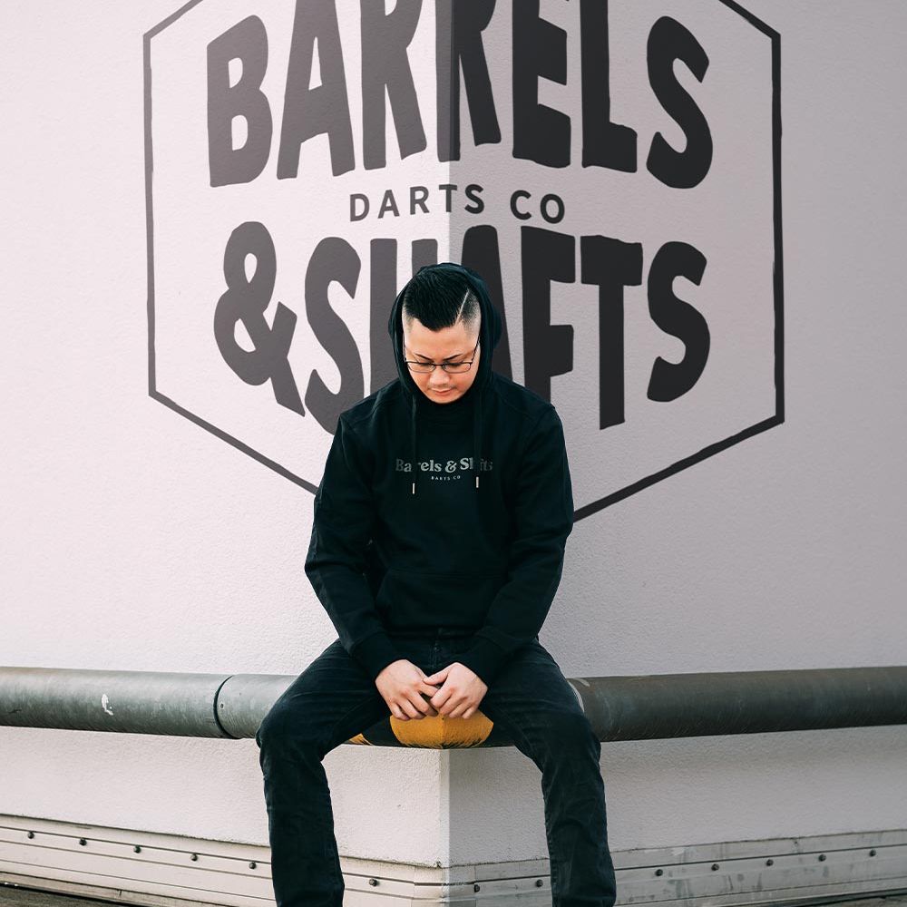 Barrels and Shafts Hoodie - Black Eine Person trägt einen schwarzen "Barrels and Shafts" Hoodie und sitzt auf einer Bank. Im Hintergrund ist ein großes Wandlogo mit der Aufschrift "Barrels & Shafts Darts Co" zu sehen.
