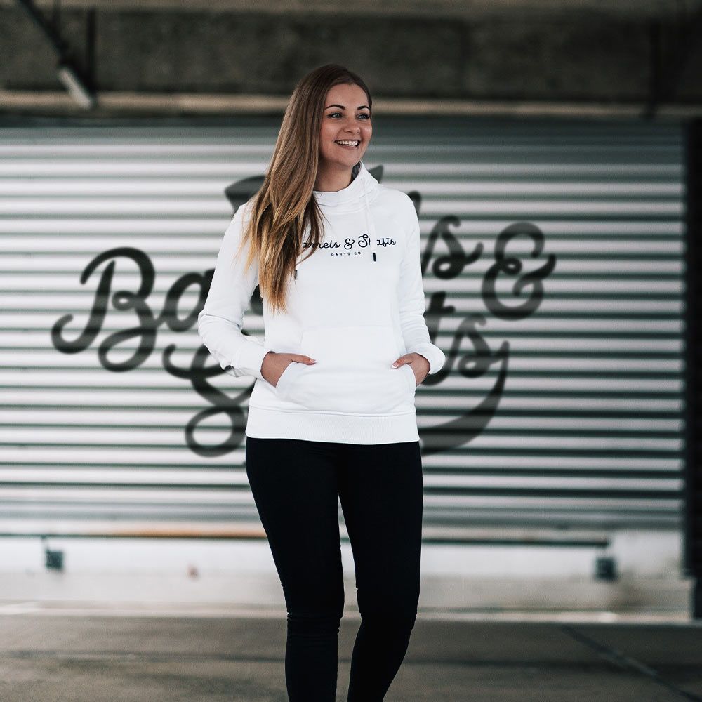 Barrels and Shafts Women's Hoodie - White Eine Frau trägt einen weißen "Barrels and Shafts" Damen Hoodie. Sie steht vor einem Hintergrund mit großem Schriftzug und lächelt.