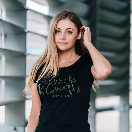Barrels and Shafts Women's T-Shirt - Black Eine Frau trägt ein schwarzes T-Shirt mit der Aufschrift „Barrels and Shafts Darts Co“. Sie steht vor einem modernen, lichtdurchfluteten Hintergrund.