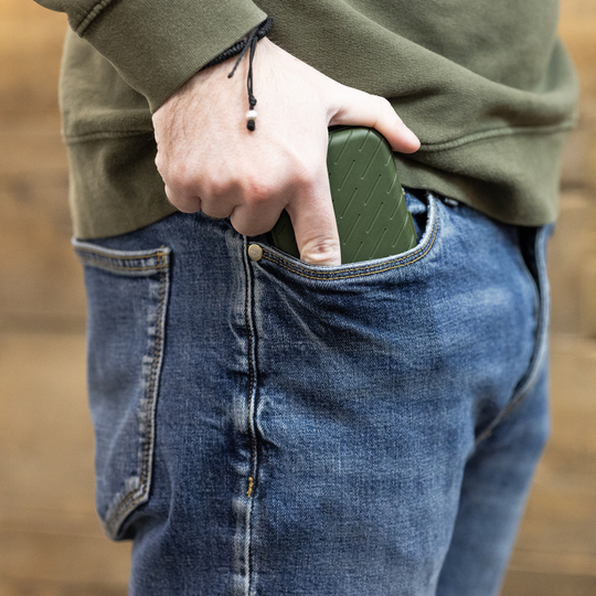 Harrows Slimcase Dart Case Eine Person steckt ein grünes Etui in die vordere Hosentasche einer Jeans. Das Etui hat eine strukturierte Oberfläche und passt gut in die Tasche.