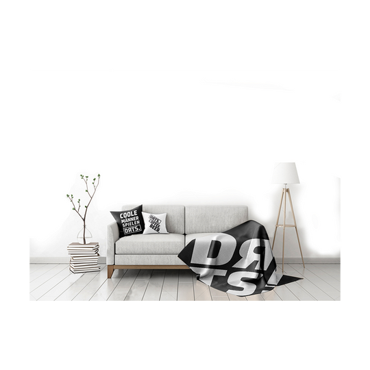 DRTS_K1_DRTS_Kissen_Schwarz_3 Das Bild zeigt ein modern eingerichtetes Wohnzimmer mit einem hellgrauen Sofa. Auf dem Sofa liegt das Produkt "SPORT1 DRTS. Kissen" mit auffälligem schwarzen und weißen Schriftzug.